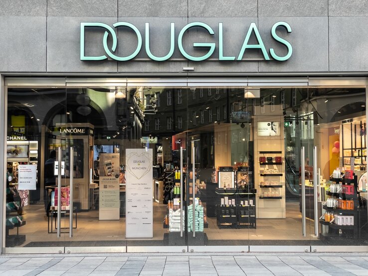 Eingang des Douglas-Geschäfts mit leuchtendem Logo, umgeben von Kosmetik-Displays und Werbung.